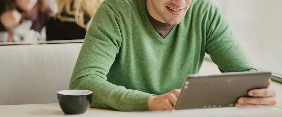 Hombre de sueter verde consulta novedades en su tablet mientras disfruta un café