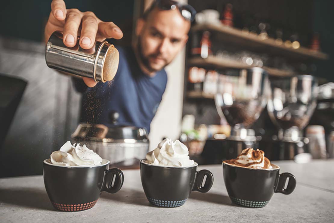 Barista espolvorea cacao sobre tazas de café con nata montada en el Día Mundial del Cacao