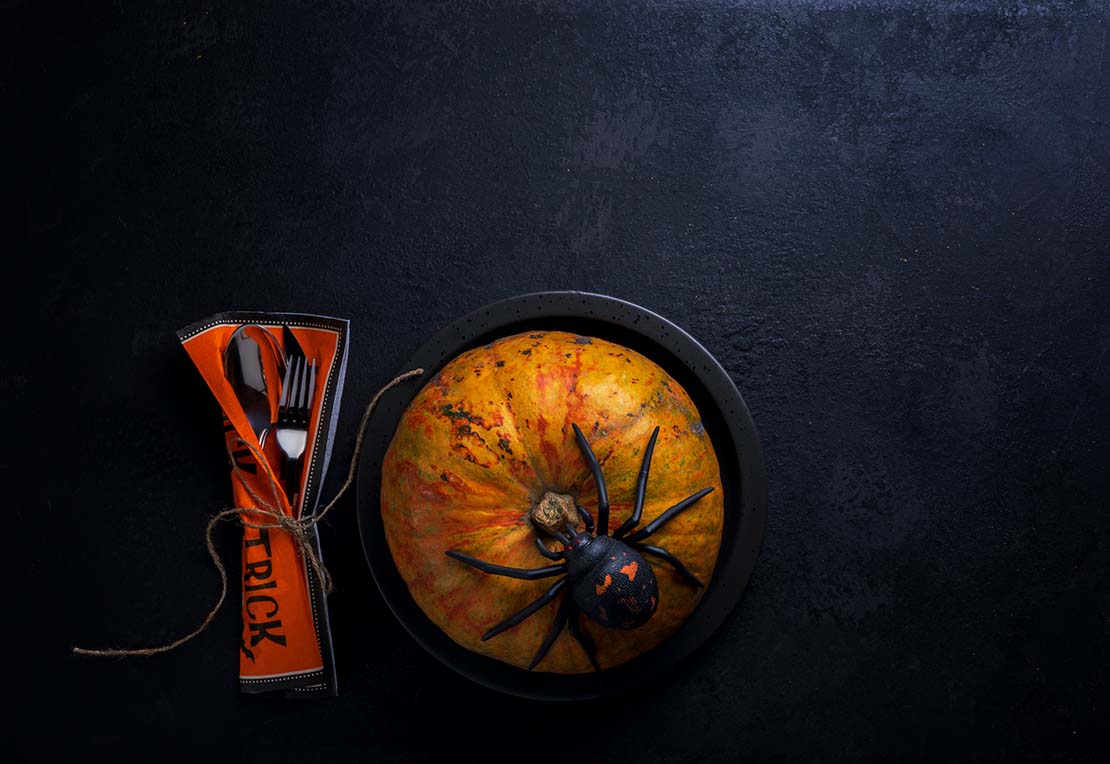 Calabaza entera servida en un tazón negro sobre la cual hay una araña de utilería y un par de cubiertos al lado