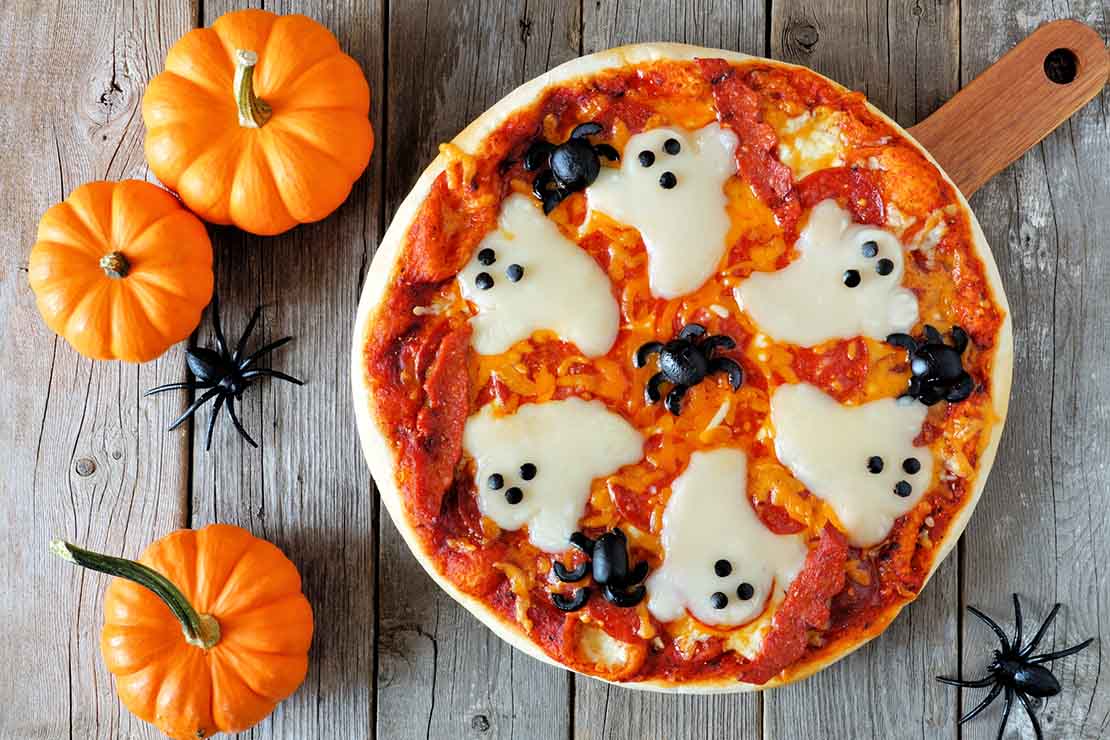 Pizza decorada con fantasmas y arañas junto a tres calabazas para ser servida en una fiesta de Halloween