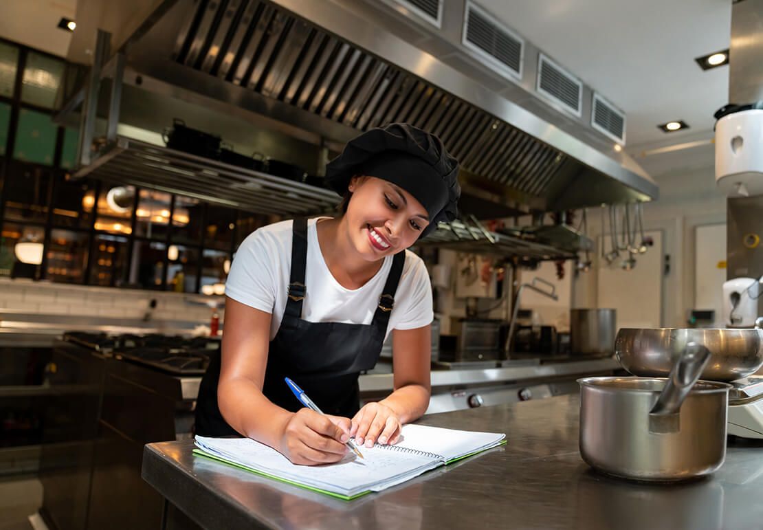 En una cocina comercial, joven estudiante del programa YOCUTA toma notas en su libreta sobre una de las mesas