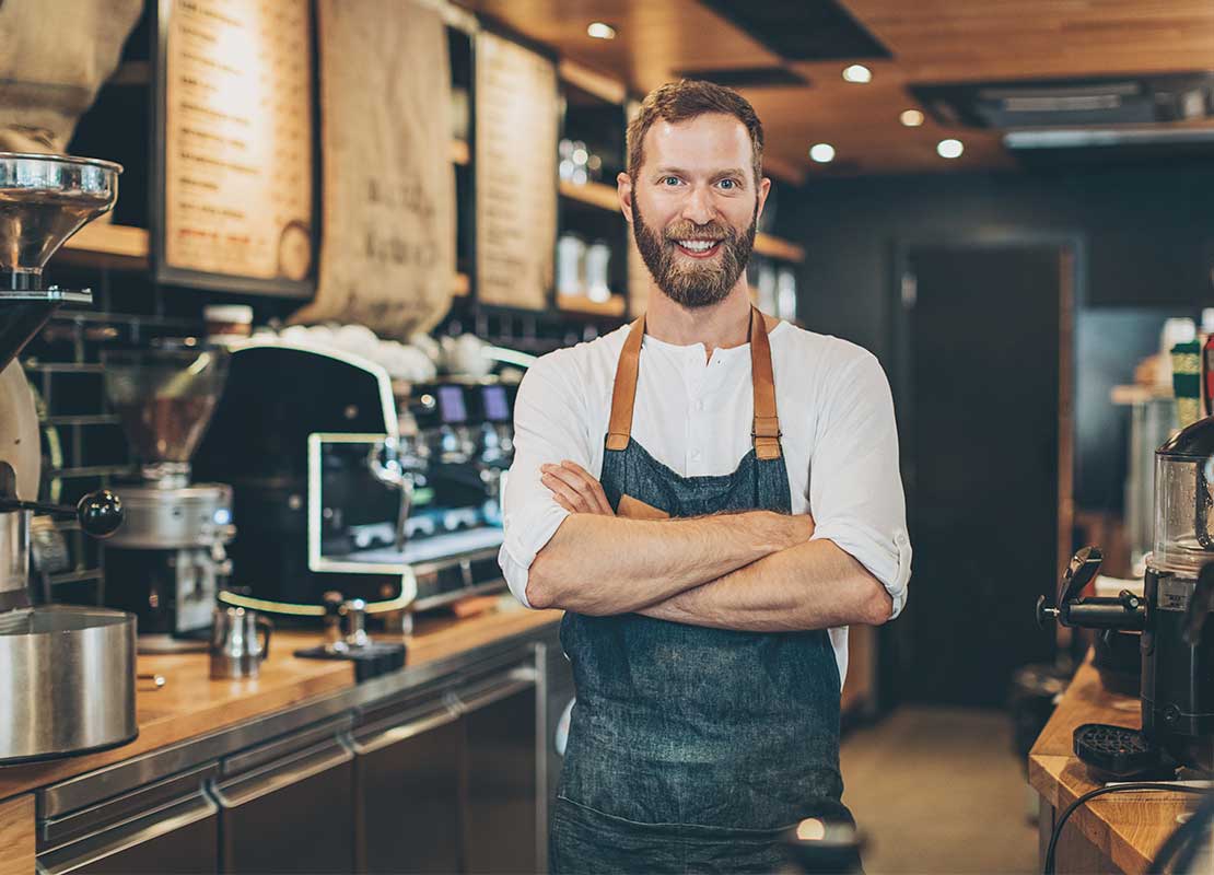 Barista cruzado de brazos tiene al fondo cocina de cafetería con máquinas y equipos de alta tecnología para hacer café frío
