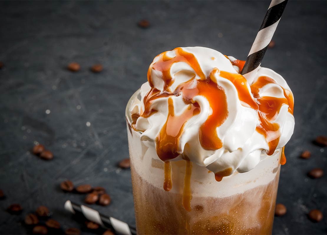 Café frío tipo frappé con crema batida y jarabe de caramelo, pajitas y granos de café en una mesa de piedra gris