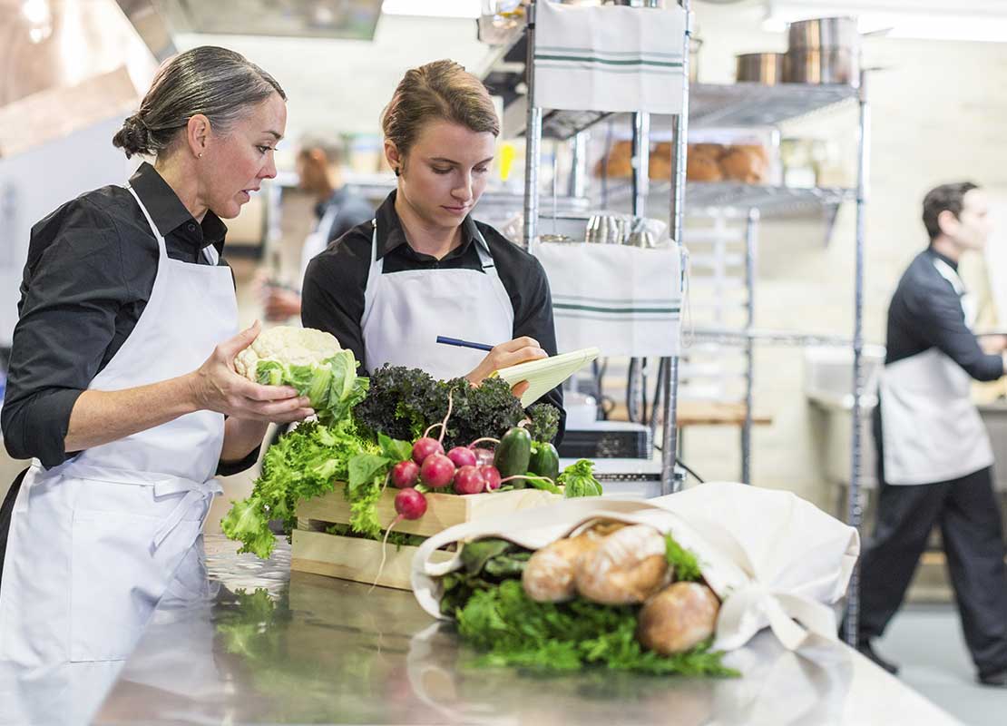 Chef ejecutiva frente a una mesa con ingredientes frescos verifica la lista de insumos recibidos que le muestra una compañera