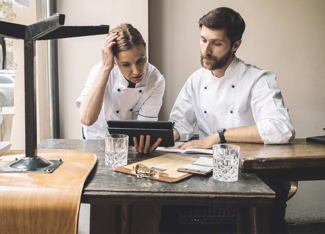 Dos chefs ejecutivos sentados a la mesa de un restaurante revisan en una Tablet el menú, platillos y presupuesto