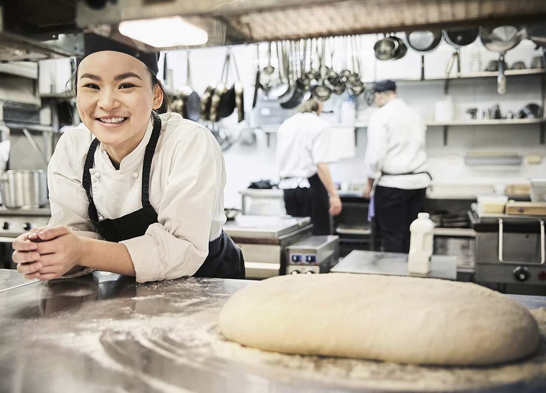Una chef inclinada sobre una mesa junto a una masa y en el fondo cocineros del grupo usando el equipamiento de cocina