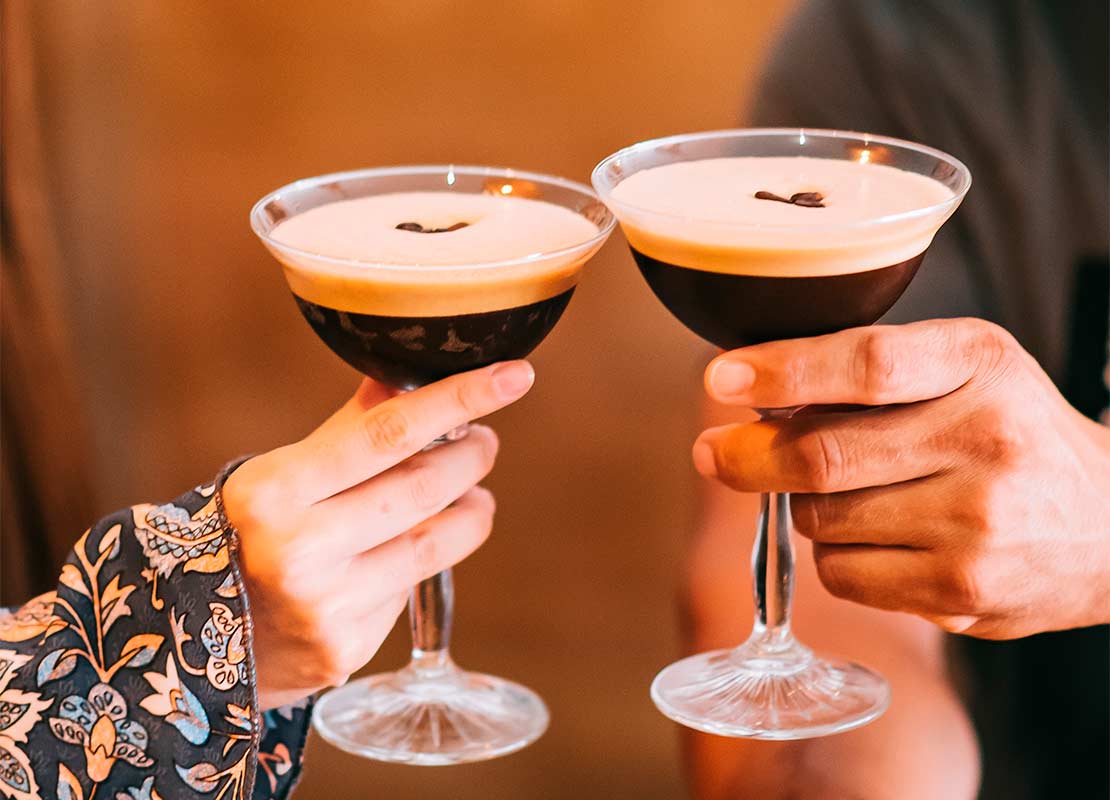 Manos sostienen dos copas de Martini Espresso, que son cócteles sin alcohol decorados con granos de café