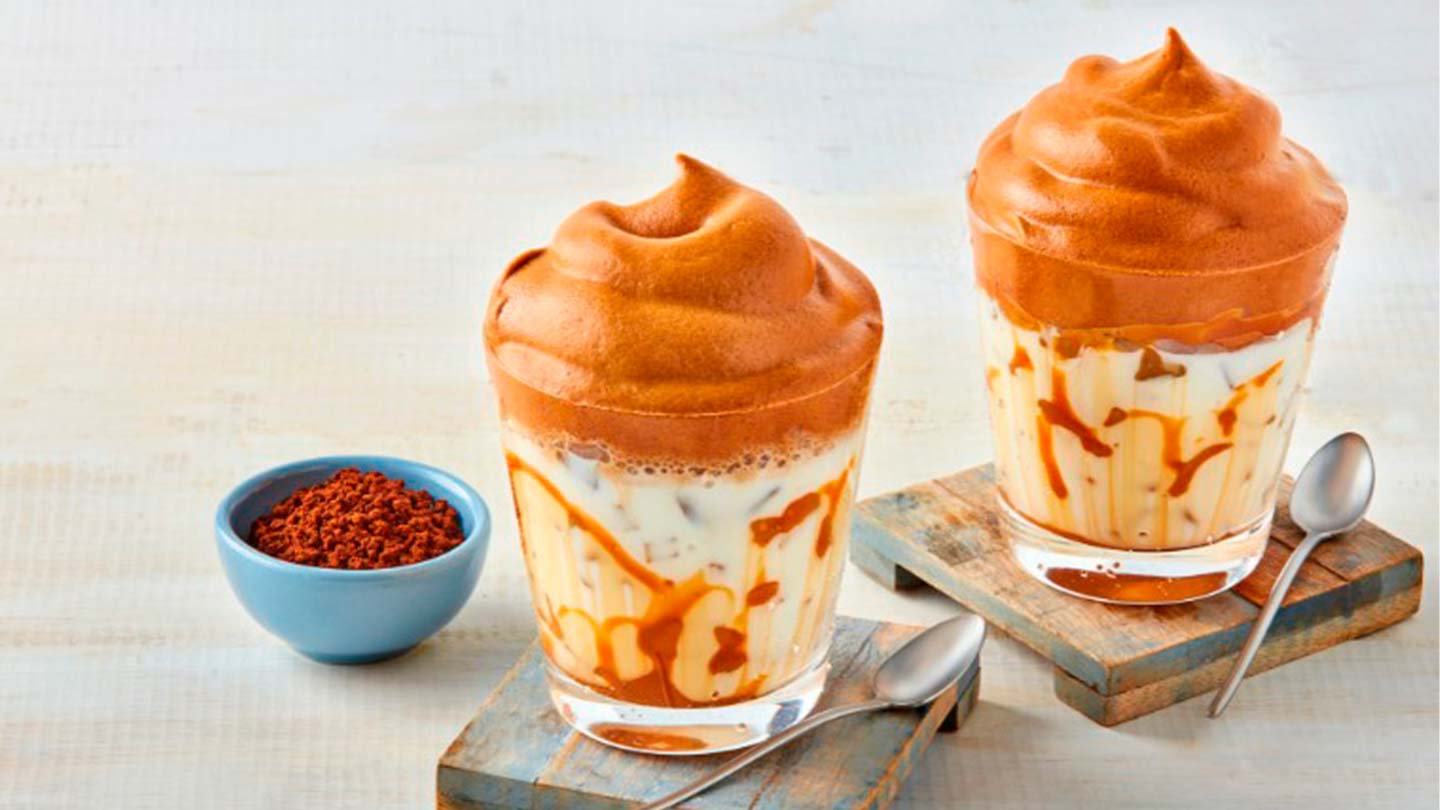 Dos vasos de café frío con hielo, leche y coronados con crema de café sobre portavasos de madera y junto dos cucharas