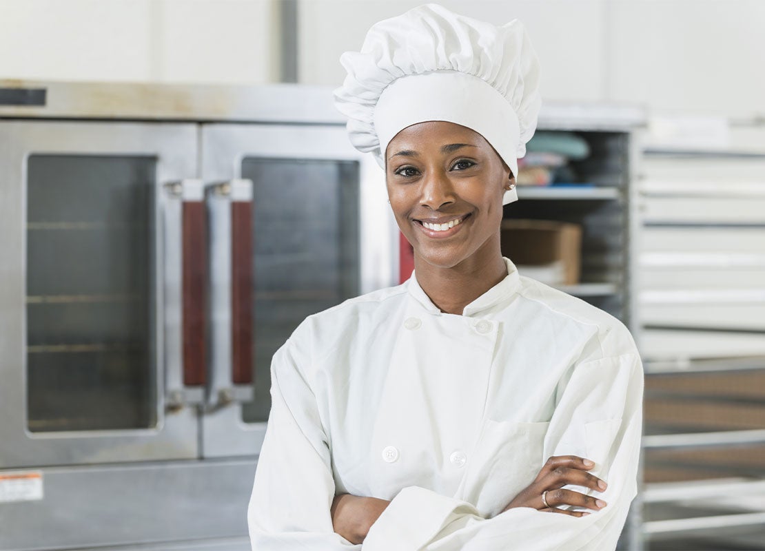 Al fondo hornos, bandejas y estanterías incluidos en un equipamiento de cocina y al frente una chef uniformada sonríe