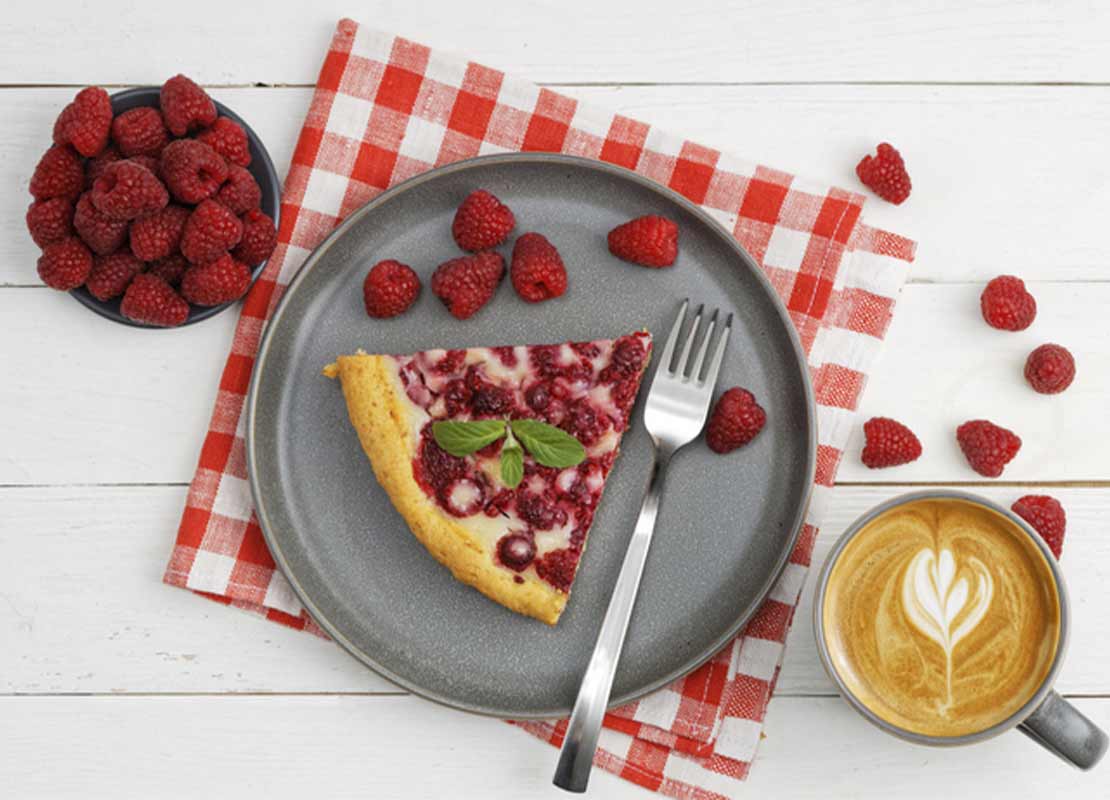 Trozo de tarta de frambuesa, una taza de café y otra de frambuesas dispuestas sobre mesa blanca para food styling