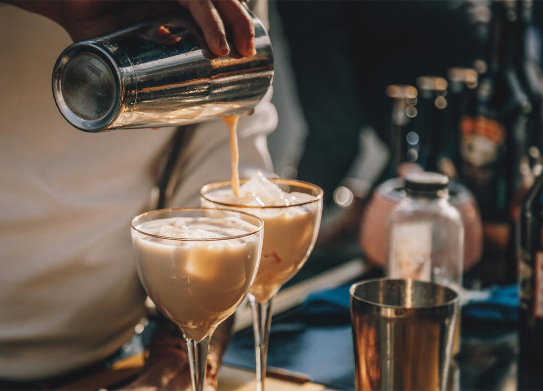 Manos de un mixólogo preparan dos cócteles sin alcohol con hielo, leche y café, lo vierte en una copa desde una coctelera