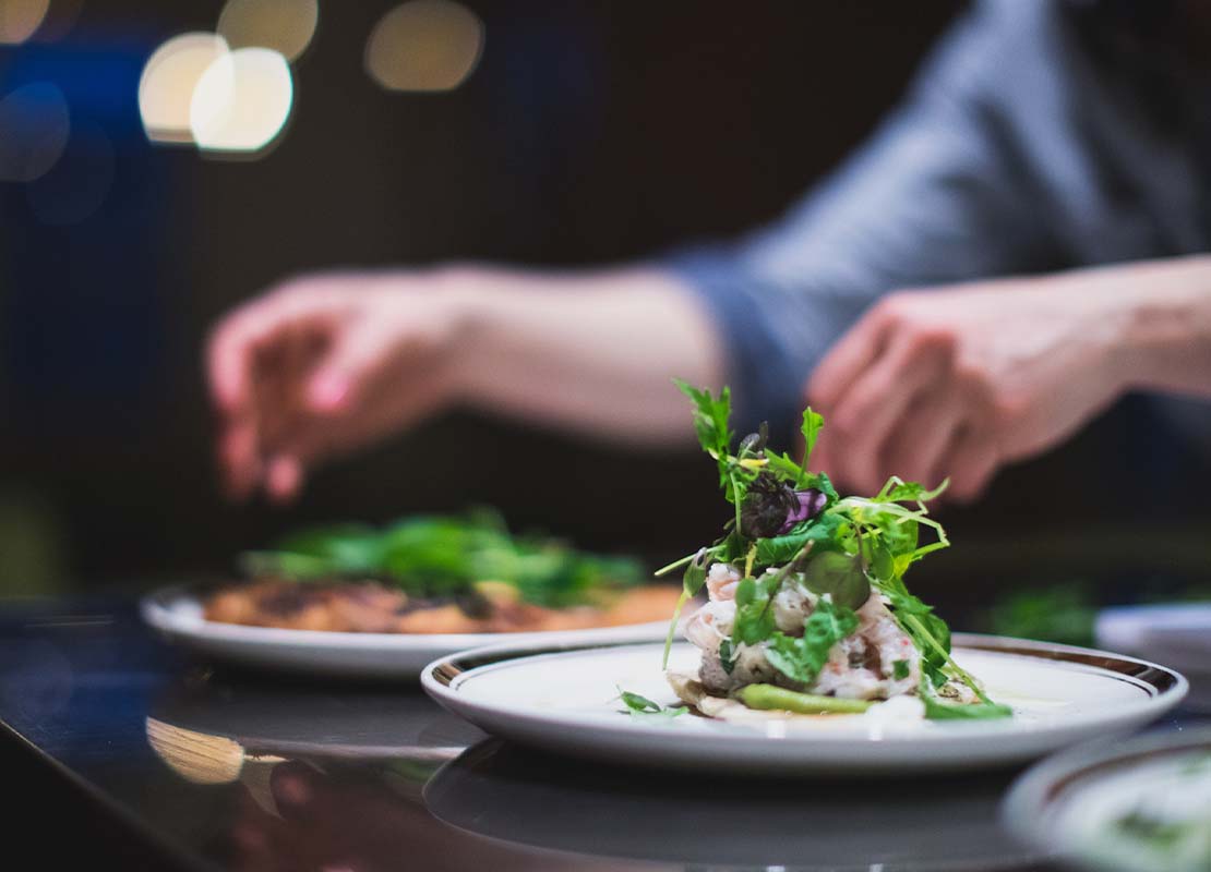 Dos platos sobre una mesa están siendo decorados y finalizados por un chef en un servicio de catering