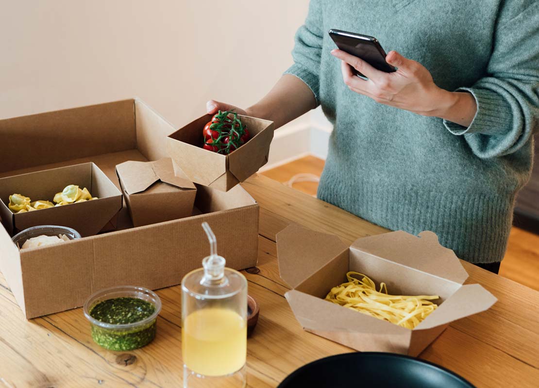 Mujer sostiene un recipiente de comida sacado de una caja de delivery mientras verifica en su celular la entrega a domicilio