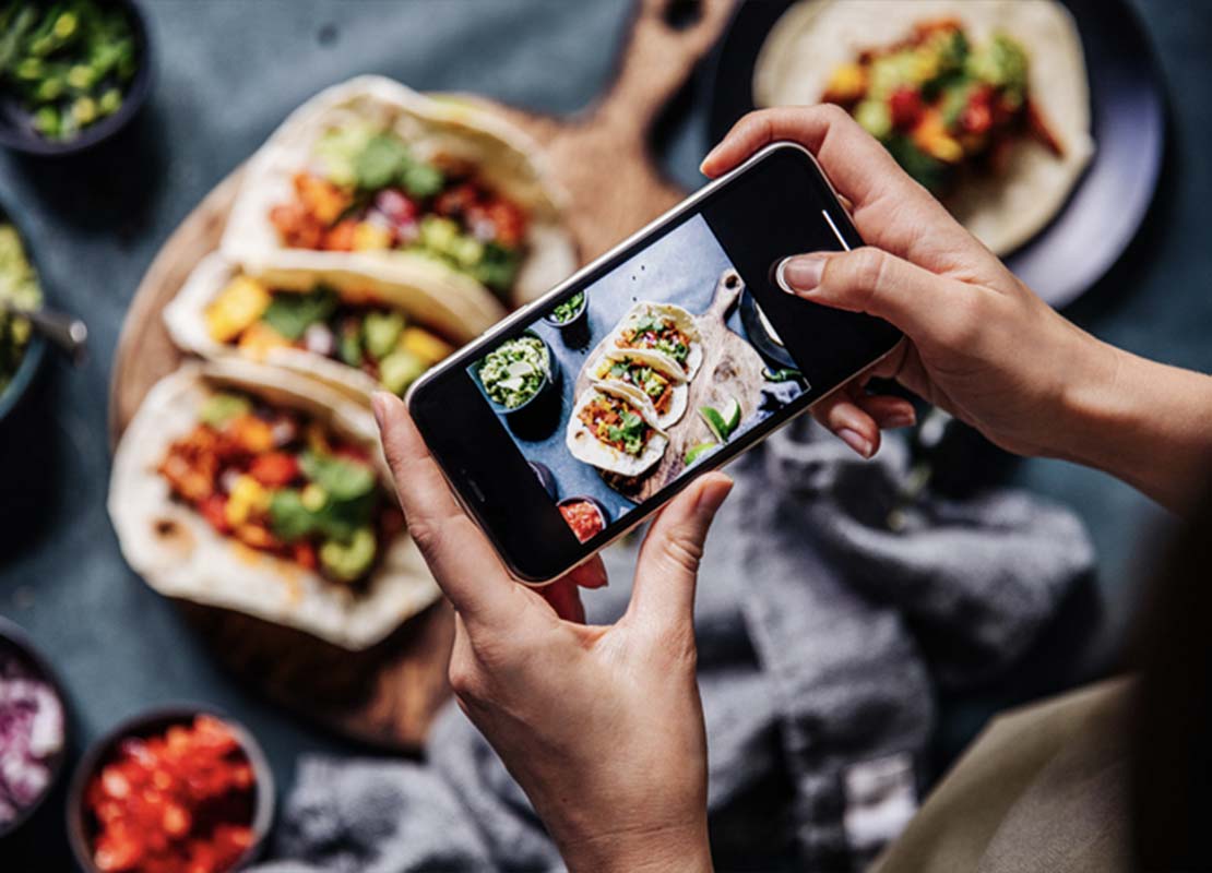 Influencer gastronómica toma fotos con su smartphone de una tabla con tacos y otros platillos de comida mexicana