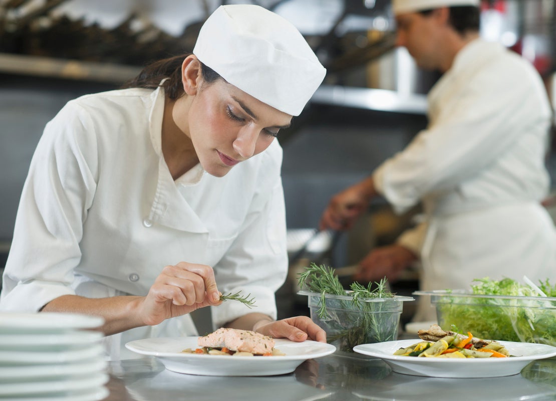 En una cocina comercial una chef decora cuidadosamente un plato con especias sobre una mesa con recipientes e ingredientes