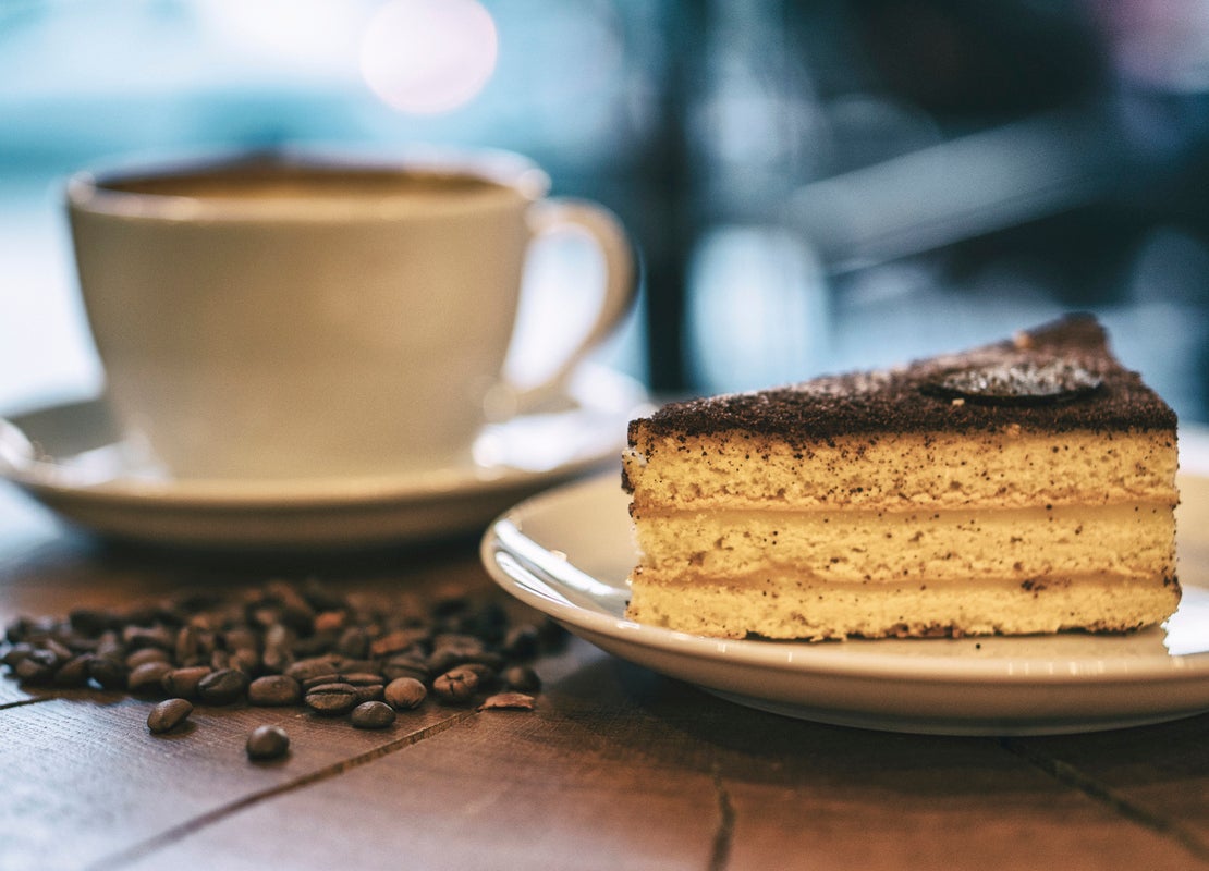 Una taza de espresso y, al frente, un trozo de tarta en una mesa de madera sobre la que hay regados granos de café tostados