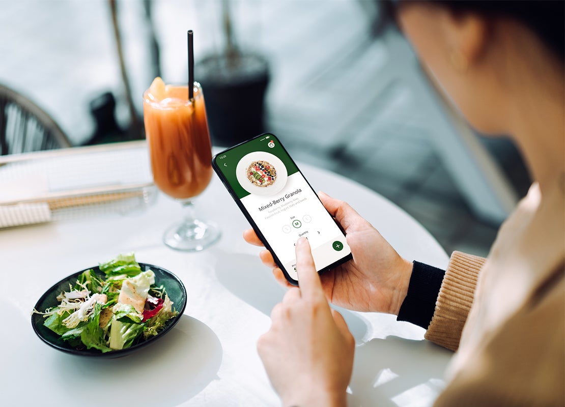 Manos de mujer sostienen un smartphone y hace autopedido de comida, frente a ella hay ensalada y una bebida sobre una mesa