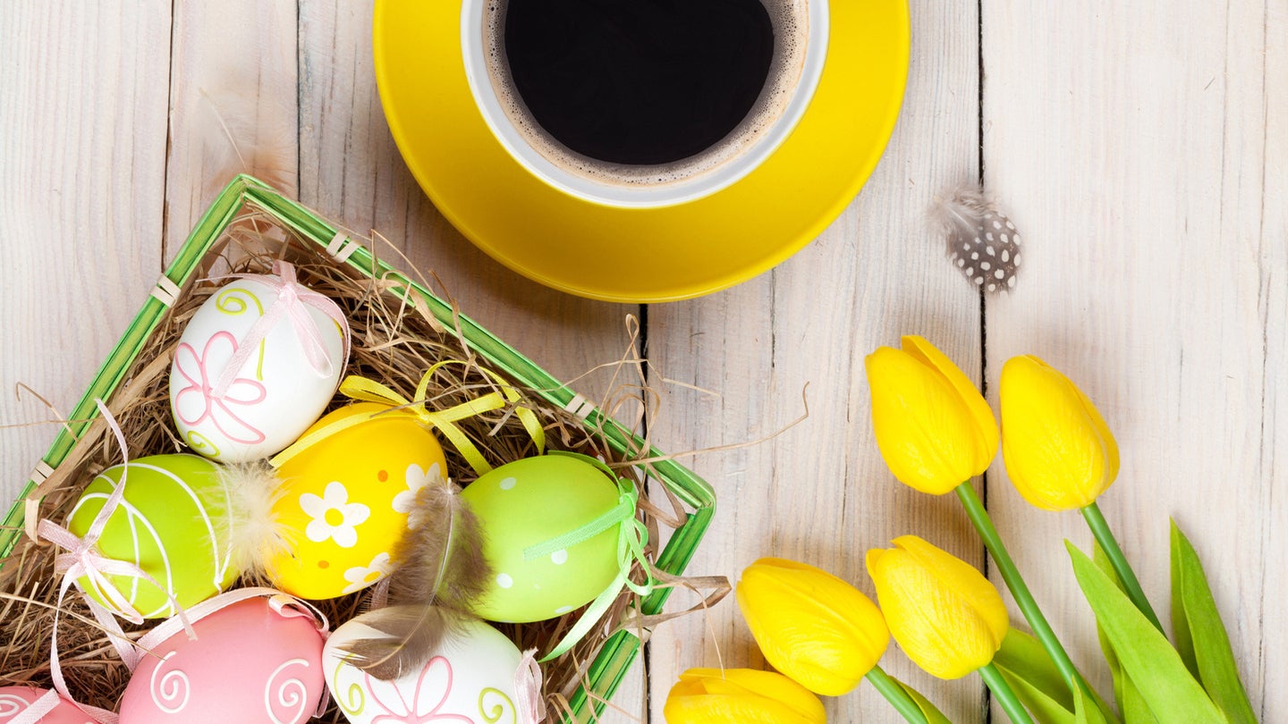 Sobre una mesa de madera 5 rosas amarillas, una taza de café y una canasta con dulces de pascua como huevos decorados