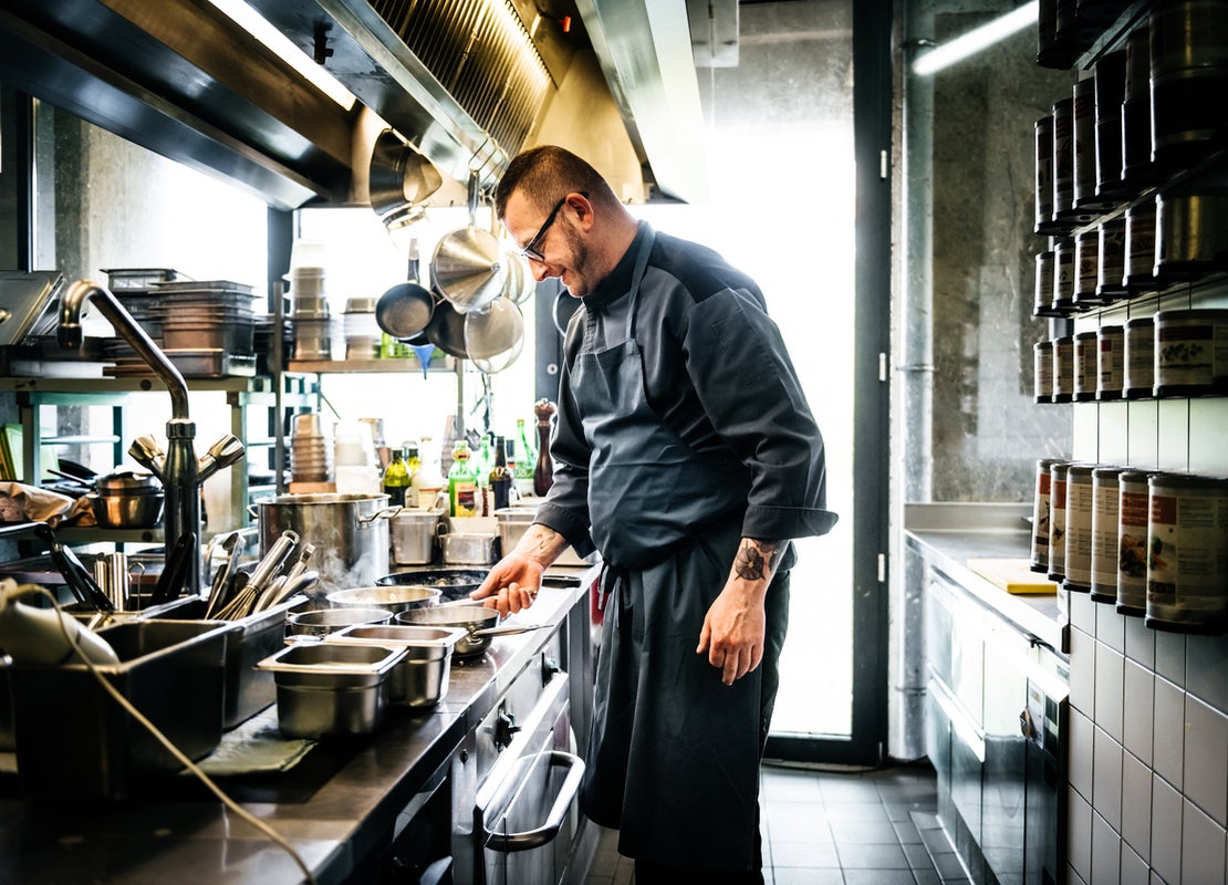 Chef revisa ollas que están sobre la estufa de una cocina de restaurante y frente a él hay utensilios en repisas y ganchos