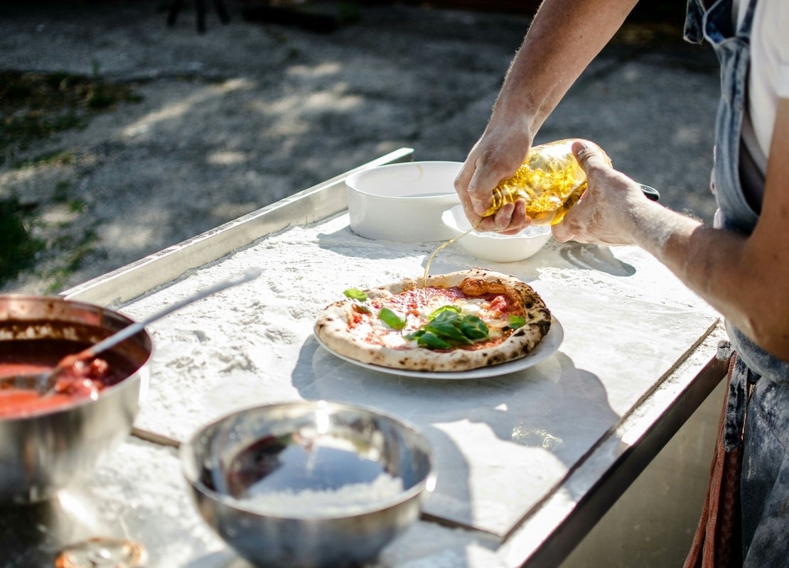 Manos de un hombre preparan al aire libre una pizza puesta sobre una mesa y alrededor recipientes con ingredientes