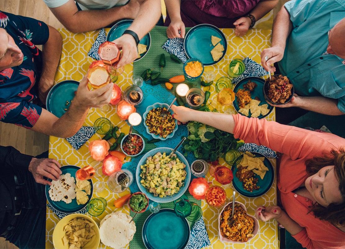 Plano cenital de una mesa con 6 comensales que disfrutan y comparten diversos platillos variados y bebidas en un restaurante