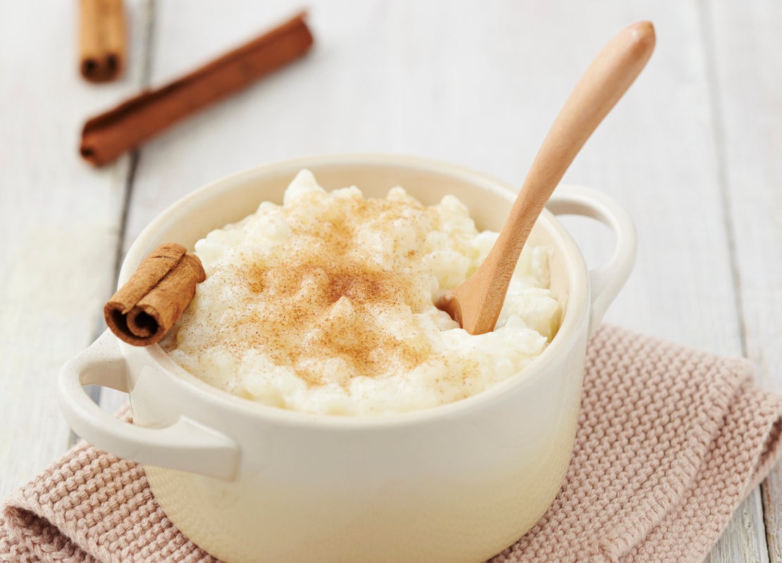 Tazón blanco de arroz con leche decorado con canela en polvo y astilla y una cuchara de madera y servido sobre una tela rosa