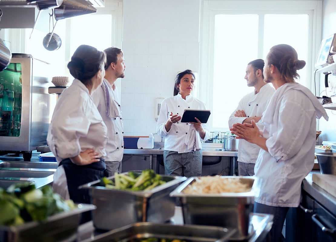 La chef de estación sostiene una Tablet en la mano mientras habla con 4 miembros de su equipo en la cocina del restaurante
