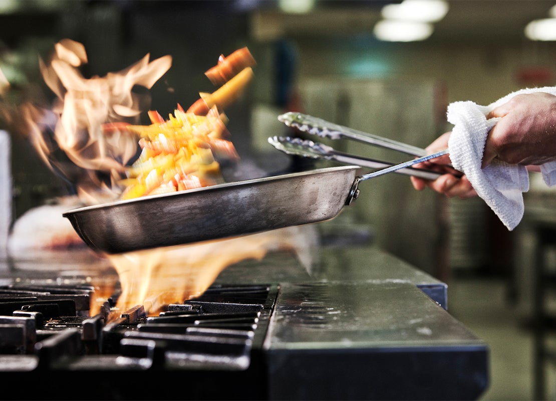 Mano de un chef sostiene unas pinzas mientras que con la otra mueve una sartén sobre la estufa para saltear los alimentos