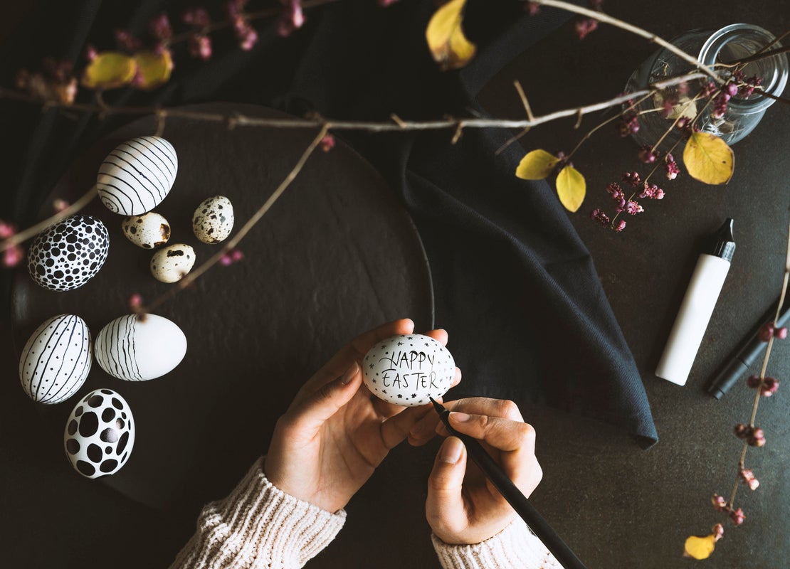 Manos pintan un huevo de pascua con marcador negro y, alrededor, otros huevos decorados y elementos para adornar