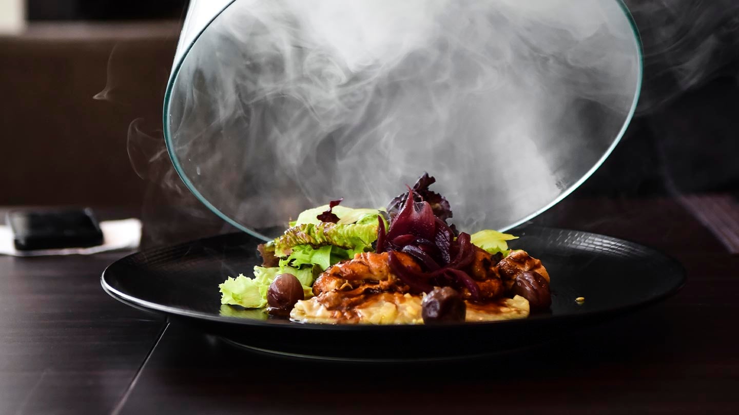 Sobre una mesa de madera se destapa un plato de comida gourmet envuelta en humo