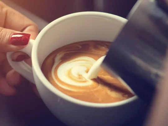 Mano de mujer sostiene una taza blanca con café mientras vierte leche con una jarra metálica para hacer latte art