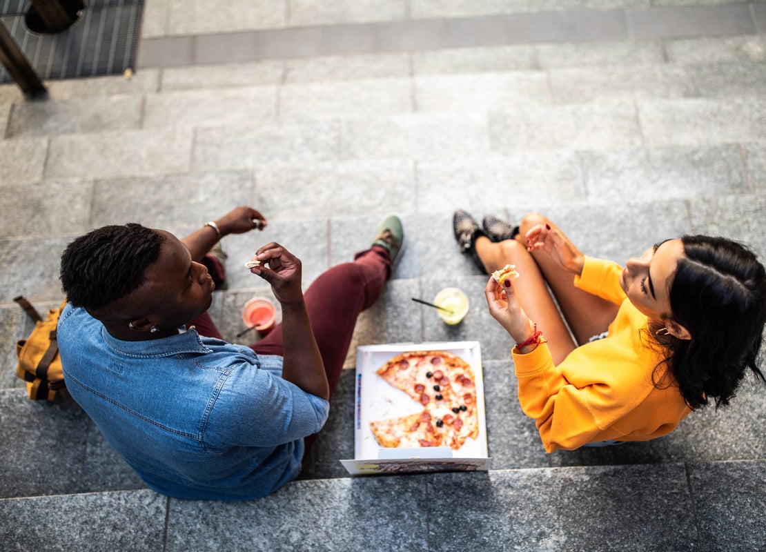 Pareja sentada en unas escaleras come pizza, mientras una caja de este alimento está en medio de ellos junto con dos bebidas