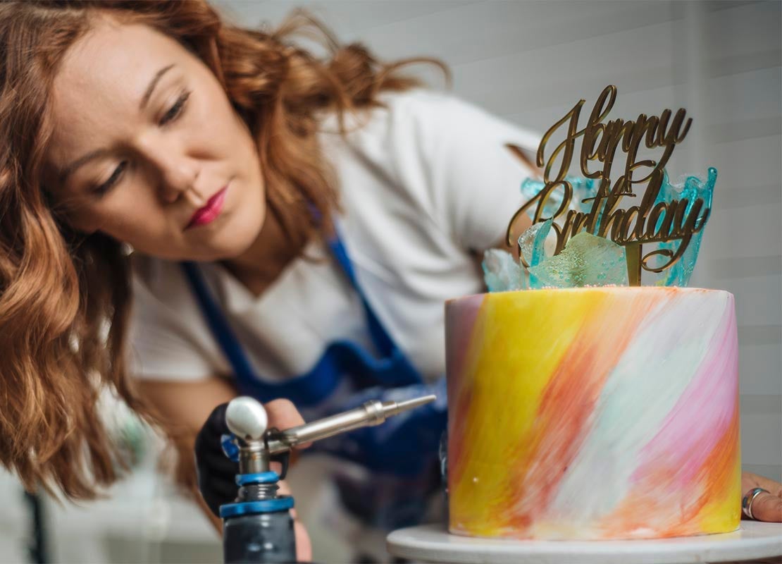 Repostera decora un pastel con aerógrafo, el cual lleva el letrero “happy birthday”