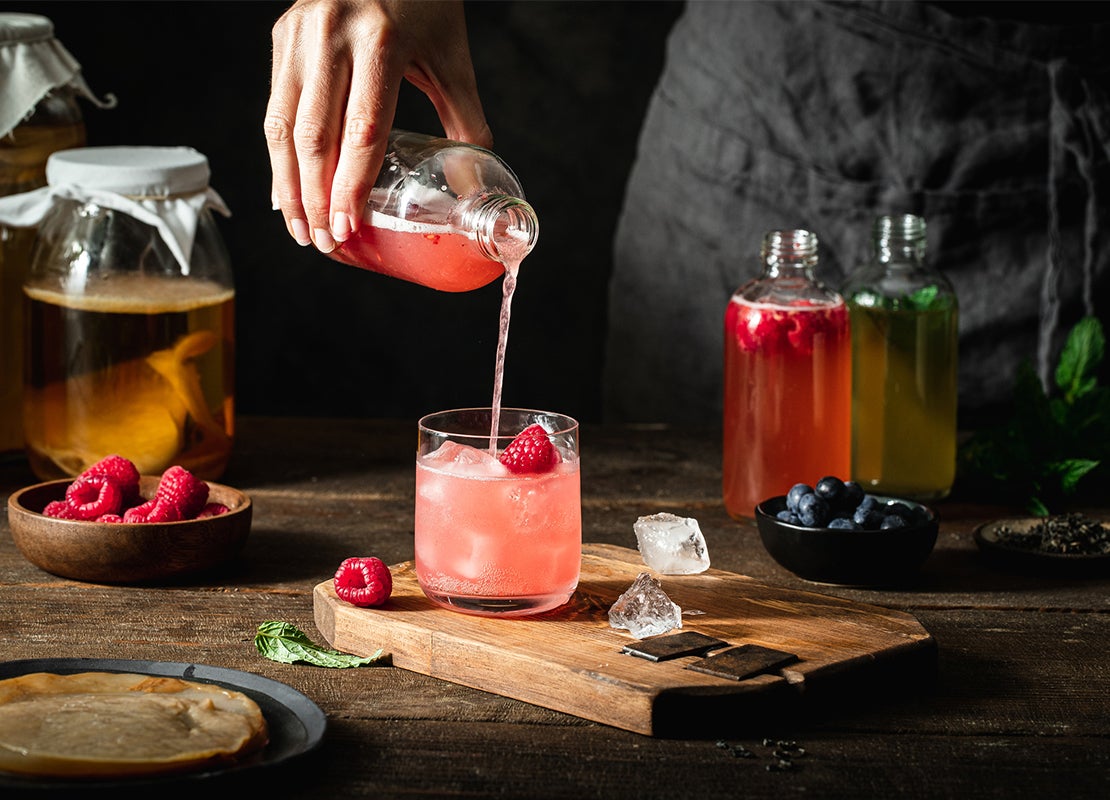Manos de mujer vierten té de frambuesa aromatizado de una botella a un vaso con hielo puesto sobre una mesa con frutas
