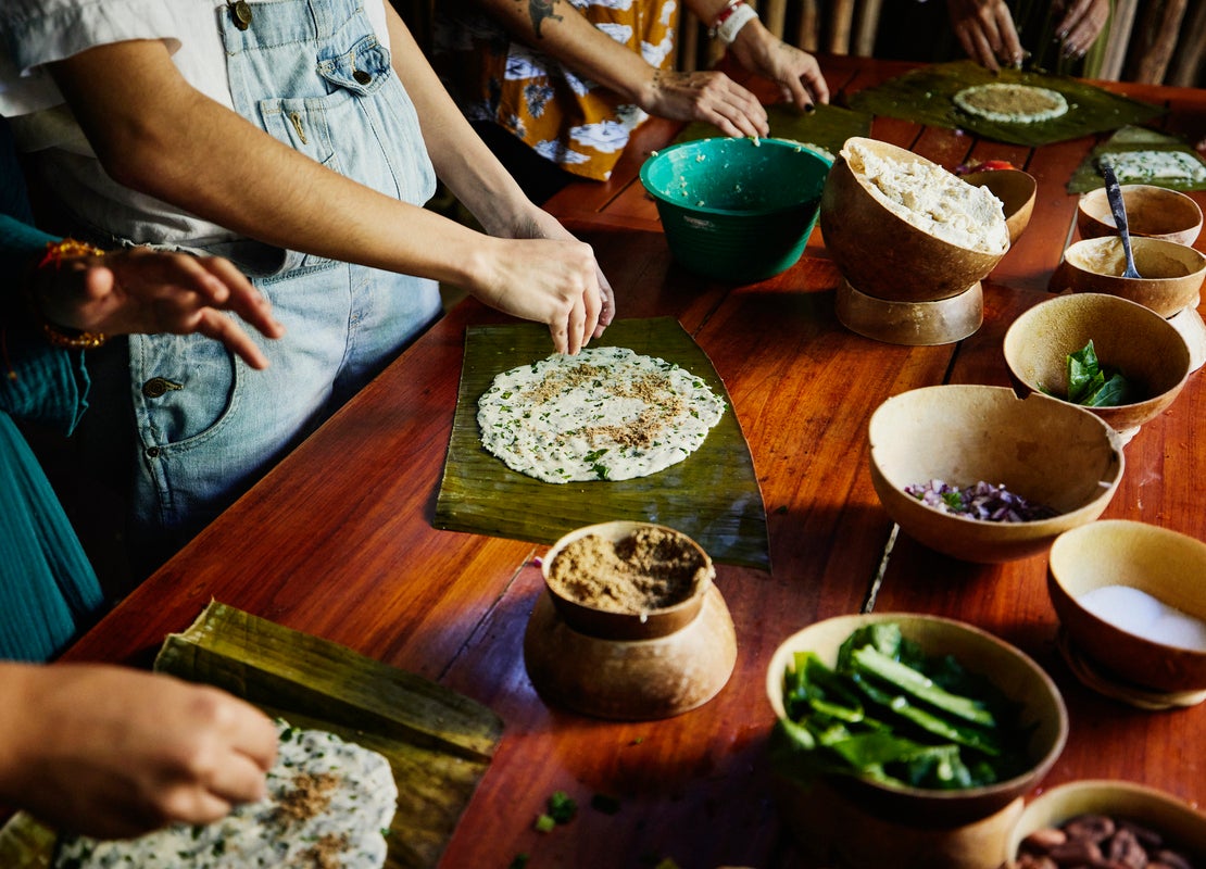 Brazos de personas preparan tamales oaxaqueños en una mesa usando hojas de plátano, masa de maíz y cuencos tradicionales