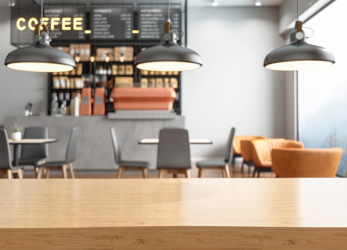 Mesa de madera vacía y al fondo el mostrador de una cafetería con productos, además de sillas y mesas cómodas