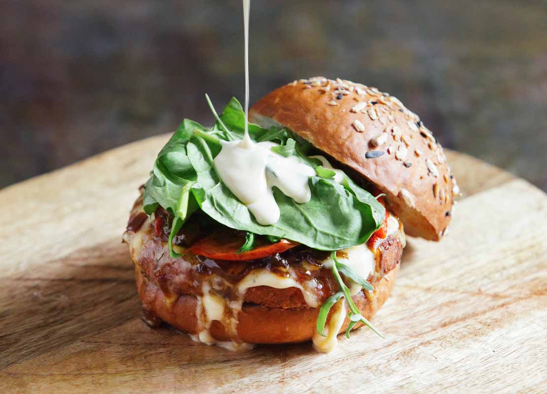 Vertido de salsa sobre los vegetales de una hamburguesa servida en una tabla