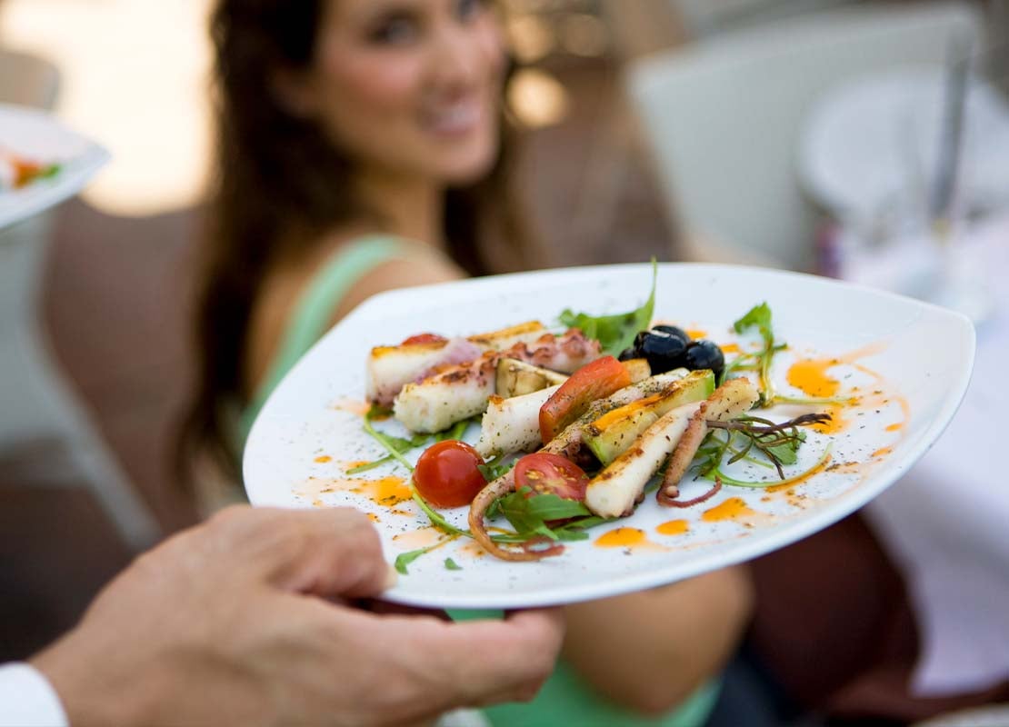 Mano de camarero sostiene un plato y al fondo está una comensal sentada a la mesa