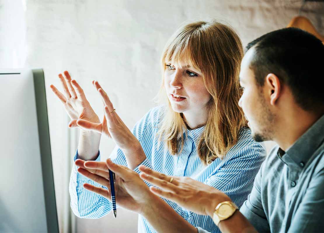 Mujer habla con su compañero mientras diseñan un estudio de mercado frente a un computador
