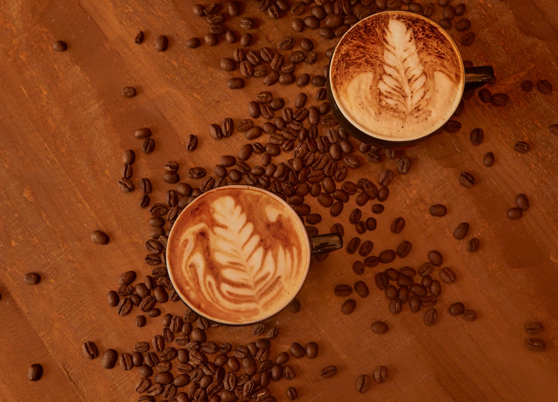 Dos tazas de café con diseño latte art rodeadas de granos tostados sobre una mesa de madera