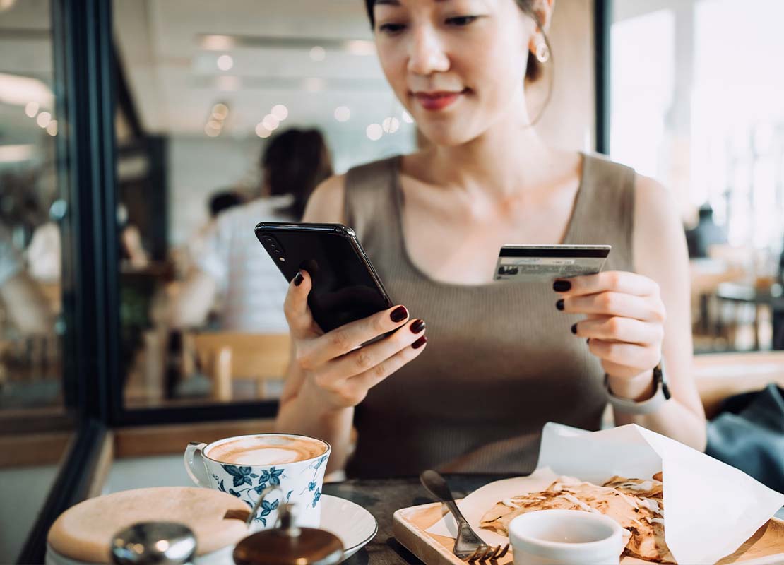 Mujer sentada en la mesa paga desde su celular su café y comida mientras sostiene su tarjeta bancaria