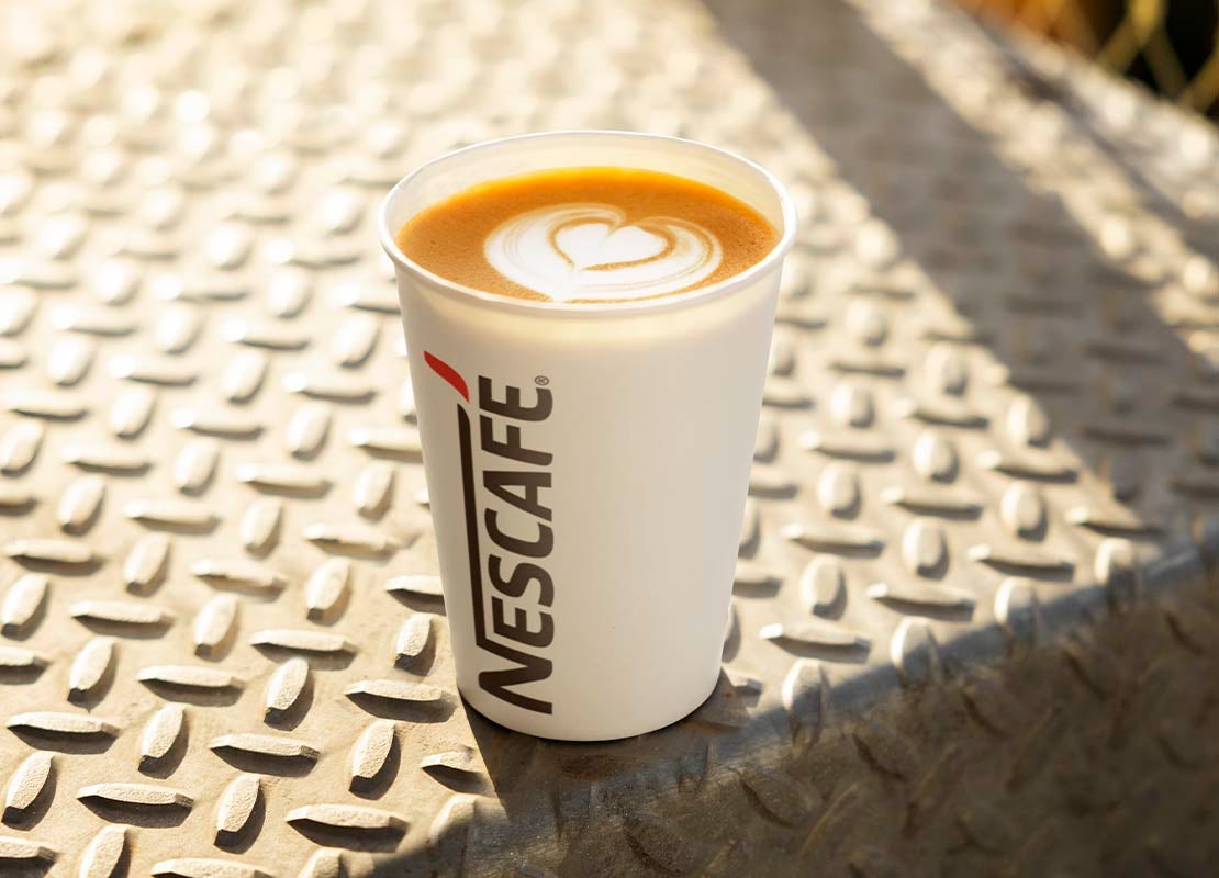 Sobre un escalón de acero, un vaso de NESCAFÉ® blanco con un café que lleva un diseño de latte art