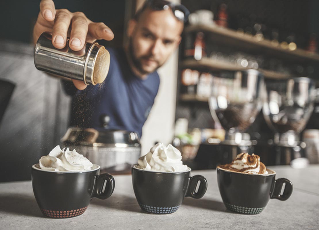 En una cafetería, un barista decora con cacao 3 tazas negras de café coronadas con crema batida