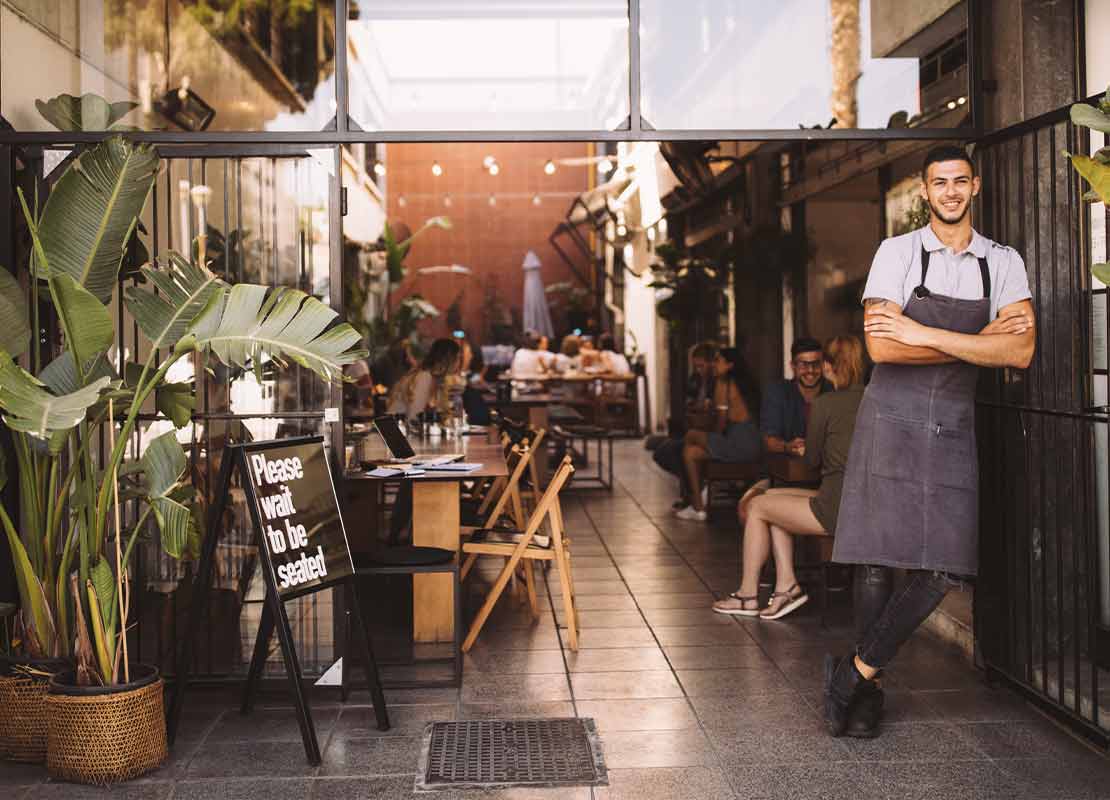 Hombre cruzado de brazos en la entrada de su negocio de comida y al fondo el mostrados y sus clientes