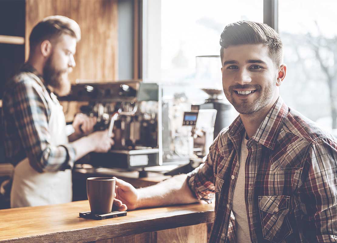 Cliente feliz sostiene una taza y al fondo el barista prepara una bebida utilizando una máquina de café