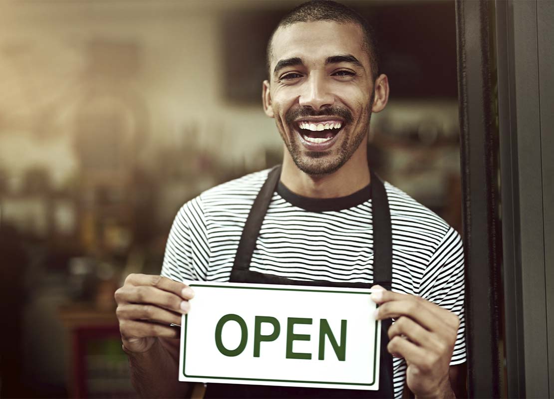Barista sonriente sostiene en sus manos un cartel de “abierto” en la puerta de una cafetería