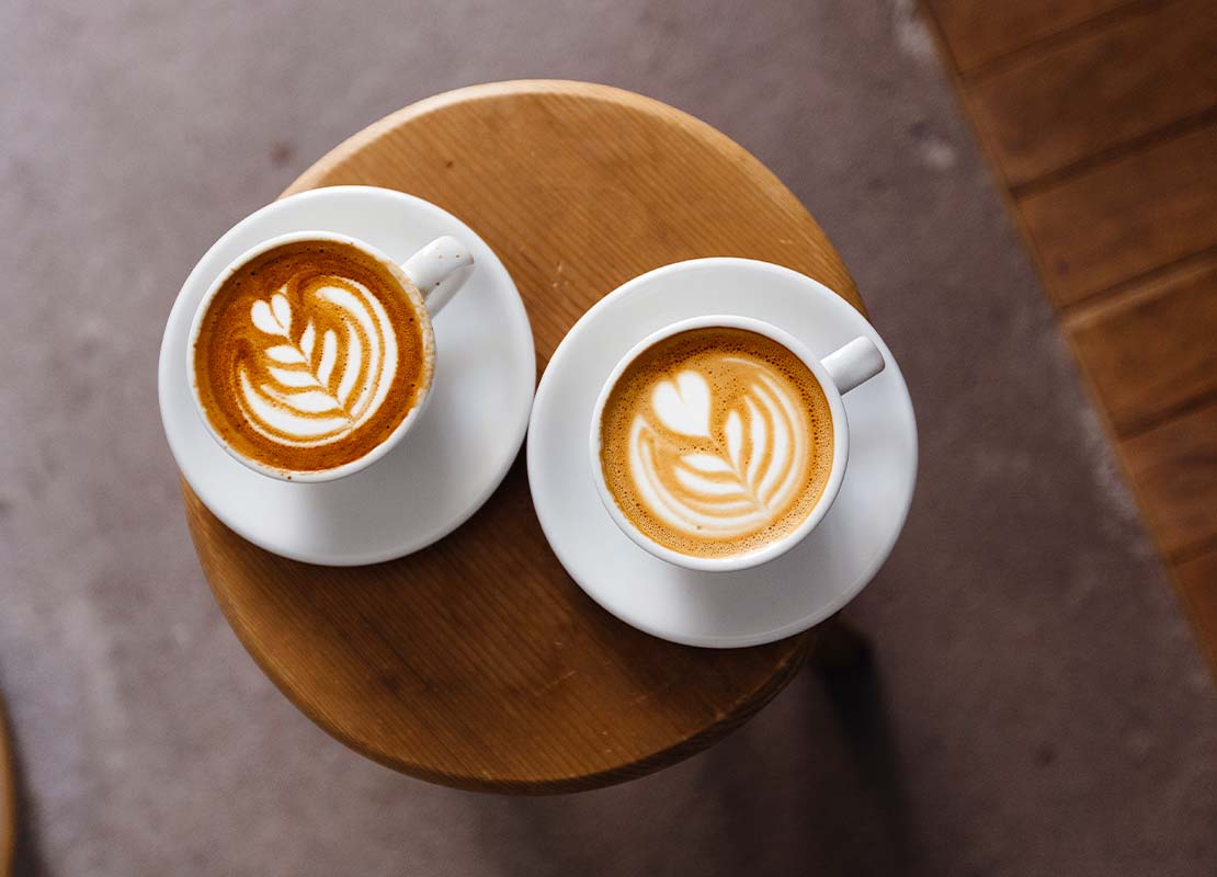 Sobre una mesa, 2 tazas de cappuccino con diseños de latte art con figuras de corazón y hojas