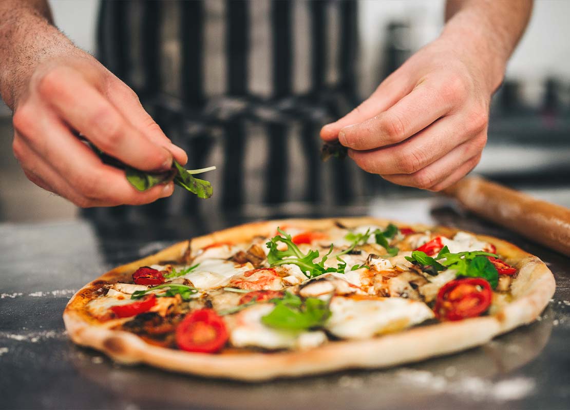 Mano de chef decorando con vegetales una pizza sobre una mesa, que hace parte del menú infantil