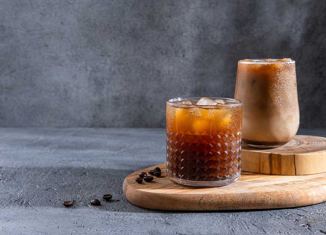 Dos vasos de cafés fríos con hielo junto a algunos granos sobre tablas