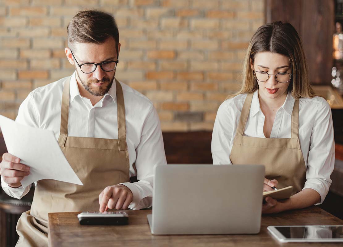 2 dueños de un café-bar sentados a la mesa con una laptop y una calculadora contabilizan y anotan